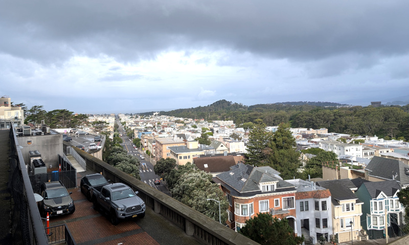 in the foreground, some cars parked on the roof of a multi-level parking garage. in the middle ground, apartment buildings and houses, seen from above. in the background, looks like a forest but is actually just Golden Gate Park, with Strawberry Hill poking up. Above, a sky of clouds with gray and white patches