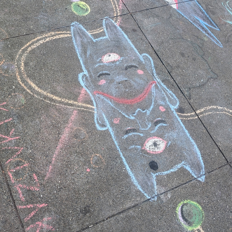 sidewalk chalk art. one of the masks. kinda demonic blue with horns. has two faces, one of which makes sense if seen right-side-up, the other upside-down. they share a mouth which frowns/smiles depending on how you look at it. each face has two closed eyes and an open third eye