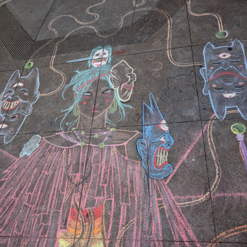sidewalk chalk art. upper part of the figure, can see most of the three demon masks and her head. also between two of the demon masks, you can see, in faint black, the remainder of the previous artwork: kind of a pudgy feline imp figure